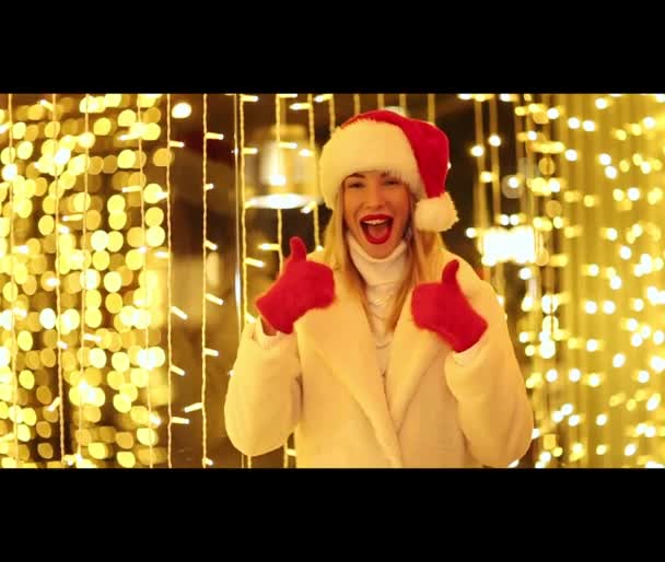 一个穿着冬衣 头戴白色皮大衣 头戴圣诞帽 头戴红色手套的漂亮而快乐的姑娘 在灿烂的灯光下翩翩起舞 面带笑容 — 图库视频影像