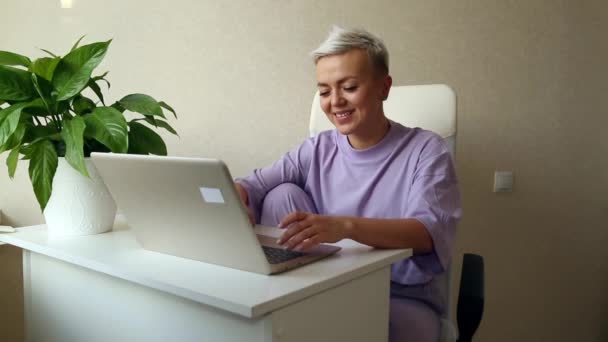 紫のパジャマに身を包んだ金髪の若い女性がノートパソコンの横に座り — ストック動画