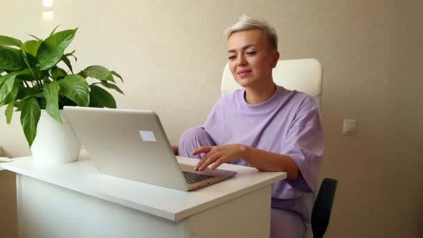 紫のパジャマに身を包んだ金髪の若い女性がノートパソコンの横に座り — ストック動画