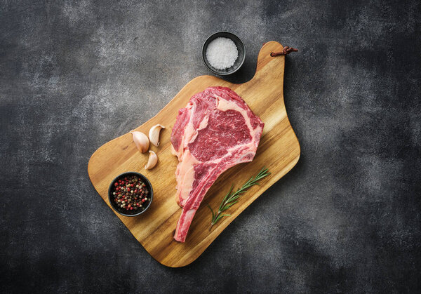 Raw Tomahawk steak on wooden board with spices for grilling, grey background