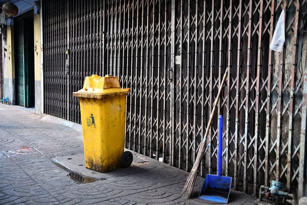 A dirty yellow plastic bin, blue powder scoop, and broomstick are in front of the sliding metal door. Bangkok street, Thailand.