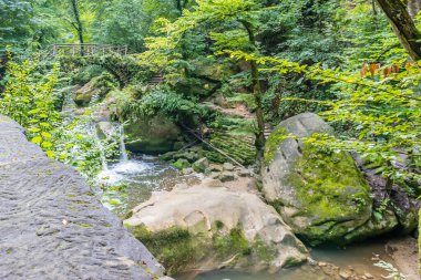 Siyah Ernz nehri ve Schiessentumpel şelalesi daha yüksek bir perspektiften görülüyor. Kayalar, merdiven ve bitki örtüsü arasında kristal berrak su akıyor. Mullerthal Patikası, Lüksemburg.