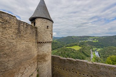 Ortaçağ Bourscheid kalesinde bir gözetleme kulesi olan dış duvar, yeşil ağaçlı vadi, bir nehir ve arka planda köy, bulutlarla kaplı bir gökyüzü, Lüksemburg