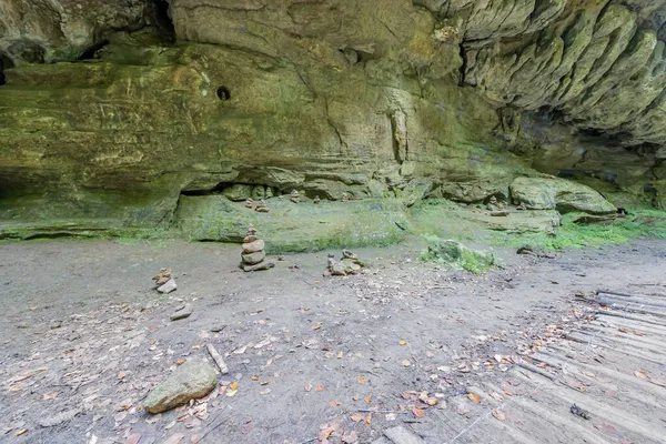 Path to huge rock formation with uneven, uneven texture and moss with cairns or apachitas at the bottom, climbing plants, summer day on the Mullerthal Trail hiking route, Luxembourg