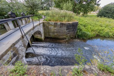 Keutelbeek stream with a small bridge over the dam and sluice with its black metal railing surrounded by green vegetation in the Sittard city park, sunny summer day in South Limburg, Netherlands