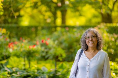 Smiling mature Mexican woman with glasses wearing white casual clothes with lush green vegetation and red flowers in the blurred background, sunny summer day to enjoy a day in park