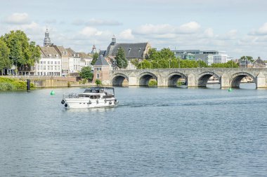 Maas river with a boat sailing, the Servatius bridge (Sint Servaasbrug) and with an urban landscape of the city of Maastricht in the background, sunny day in the South Limburg, Netherlands