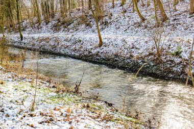 Stream with sun reflection on water surface next to snowy hill with wild vegetation in Dutch countryside, winter day in Stammenderbos, South Limburg, Netherlands