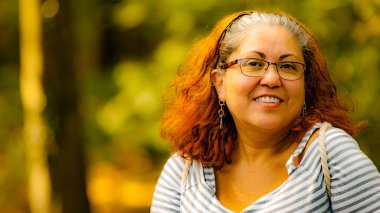 Mature Latin woman with a wide smile and looking at the camera, gray hair, glasses, casual clothes, wonderful sunny autumn day with a green and yellow blurred background. Space for text