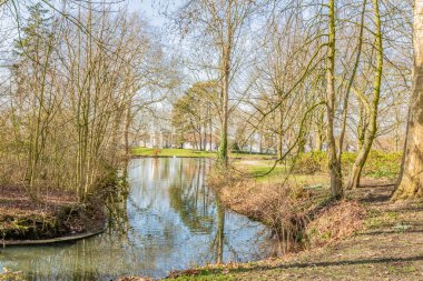 Stream with clean and crystal clear water with reflection on the water, surrounded by bare trees, green brown grass and vegetation, sunny day in Proosdij park in Meerssen, South Limburg, Netherlands