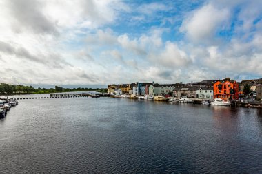 Calm waters on the River Shannon, boats moored on the shore at the docks, picturesque houses in the background, cloudy day covered by clouds in the town of Athlone, County Westmeath, Ireland
