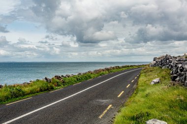 View of the rural coastal R477 road between the sea and the rocky area of the Burren, geosite and geopark, Wild Atlantic Way, cloudy spring day in county Clare, west coastal of Ireland