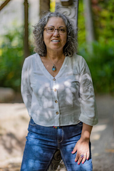 Smiling and happy Mexican mature woman with gray-black hair wearing glasses leaning on a wooden pole with green vegetation with a blurred background, enjoying a sunny summer day