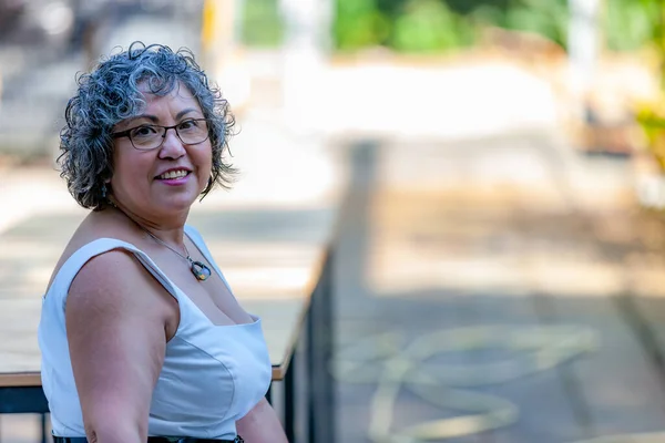 Headshot close up portrait of a mature Mexican woman smiling and looking at the camera, short gray hair, glasses and white dress with a blurred background, sunny summer day in the Netherlands