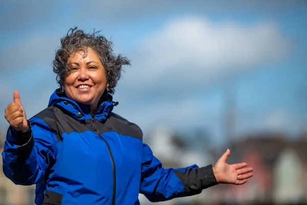 Portrait of a mature Mexican woman with arms outstretched and her hair disheveled by the wind, making a sign with her thumb raised, short gray hair, no makeup, blue jacket, Ireland. Space for text