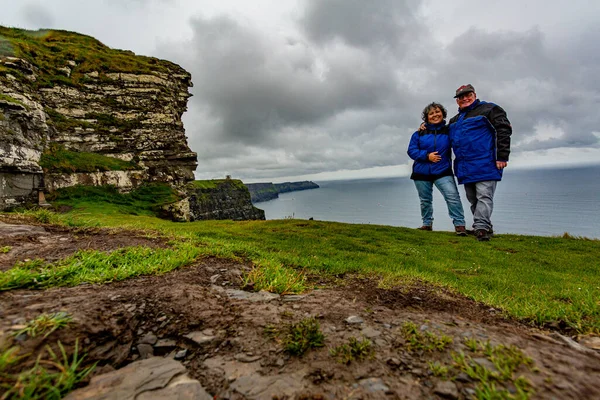 Arka planda okyanus olan Moher Kayalıkları 'nın kenarında durup kameraya, Geosites' e, Geopark 'a, Wild Atlantic Way' e bakan mavi ceketli gezginler İrlanda 'nın Clare ilçesinde harika bulutlu bir bahar günü.