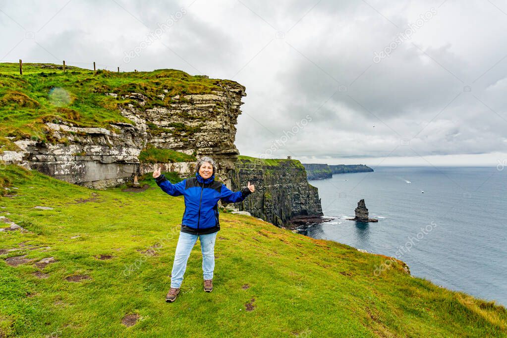 Mature Mexican woman with arms outstretched on the Cliffs of Moher, Branaunmore Sea Stack in the background, short gray hair, blue jacket, cloudy spring day in County Clare, Ireland. Wild Atlantic Way