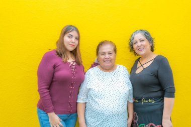 Torunu, büyükannesi ve kızı kameraya bakıyor. Üç nesil boyunca Meksikalı kadınlar sarı arka planda sıradan kıyafetler içinde gülümsüyor.