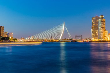 Cityscape on a calm night with its buildings and the Erasmus cable-stayed bridge over the Maas river, yellow lights, clear sky in the Netherlands Holland. Long exposure photography