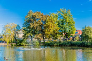 Pond with a fountain running with slight reflection in the water, lush trees with yellow green foliage and houses in the background, sunny day in early autumn in Voerendaal, South Limburg, Netherlands