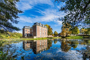 Voerendaal, South limburg / Netherlands. October 14th, 2018. Cortenbach Castle with its pond with mirror reflection and dry leaves on the water surrounded by trees, sunny autumn day with a blue sky