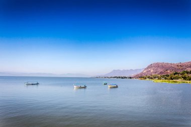 Boats anchored on the calm waters of Lake Chapala with mountains in the background blurred and misty, sunny day with a blue sky in the state of Jalisco Mexico
