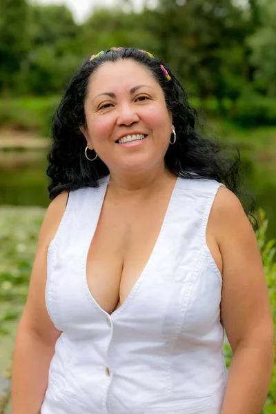 Middle-aged Latin woman with a wide smile and looking at the camera, long black hair, no makeup, low-cut white blouse and colorful headband with blurred green background, sunny spring day in a park