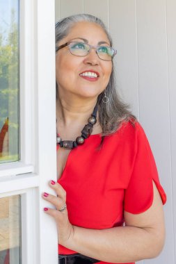 Mutlu olgun bir kadın evinin girişinde dikilip, kırmızı bir elbise içinde düşünceli ve yansıtıcı, bronz tenli, gri saçlı, gözlüklü, siyah aksesuarlı, açık renk makyajlı.