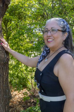 Siyah bir yaz elbisesi, bronz ten, gri saç, saç bandı, gözlük, beyaz bluz, açık makyajlı mavi etek, güneşli bir günde poz veren olgun bir kadın.