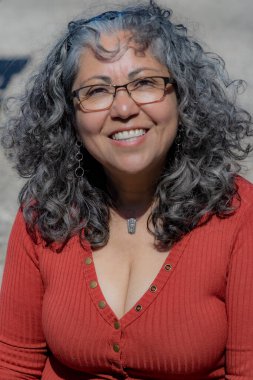 Geniş tebessümlü ve kameraya bakan olgun bir Latin kadın, bronzlaşmış ten, gri saç dalgalı ve rüzgarla dağılmış, saç bandı, gözlük, kahverengi bluz, makyajsız, günlük tarz, bulanık arka plan