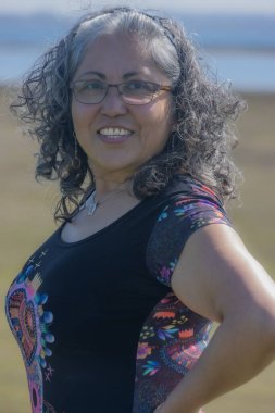 Geniş tebessümlü ve kameraya bakan neşeli olgun Latin kadın, rüzgarın savurduğu gri dalgalı saçlar, bronzlaşmış ten, gözlük, saç bandı, siyah bluz, günlük tarz ve makyajsız, bulanık arka plan.