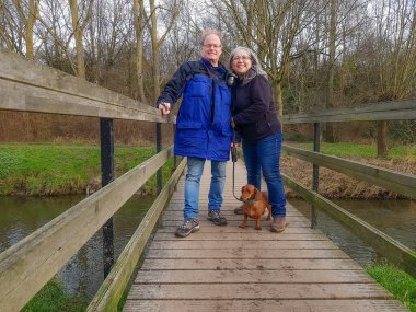 Olgun yürüyüşçüler kahverengi kısa saçlı daksundlarıyla bir derenin üzerindeki ahşap köprüde dikiliyorlar, kameraya bakıyorlar, sisli ve bulanık arka planda çıplak ağaçlar, doğa rezervi, Hollanda