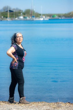 Olgun Latin kadın Grevelingen Gölü kıyısında poz veriyor, kameraya bakıyor, arka planda demirli yelkenliler, siyah kıyafetler, bronzlaşmış ten, Zonnemaire, Zeeland, Hollanda 'da güneşli bir gün.