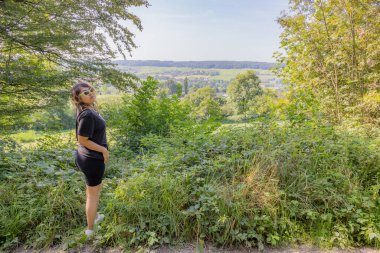 Siyah spor kıyafetli genç bir kadın bir tepede dikilip kameraya bakıyor. Arka planda yeşil ağaçlar olan Hollanda manzarası. Vijlen, Güney Limburg, Hollanda 'da güneşli bir gün.