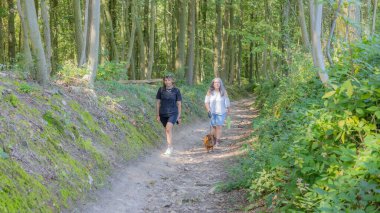 Dachshund, yeşil bitki örtüsü ve arka planda ağaçlarla yürüyüş yolunda sessizce yürüyen iki Latin kadın, gözlük, şort, günlük tarz, Vijlen, Güney Limburg, Hollanda 'da güneşli bir gün.