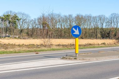 Kırsal yol, yuvarlak mavi bir plaka ve beyaz bir ok ile yuvarlak mavi bir levha ile: sürüş yönü, bulanık arka planda bol çıplak ağaçlar, Hollanda 'da güneşli bir gün