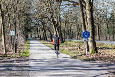 Someren, North Brabant, Netherlands. February 27, 2022. Cyclist pedaling on a bike lane, a countryside road, sign indicating: bike and moped path, routes 11, bare trees, back to the camera, sunny day clipart