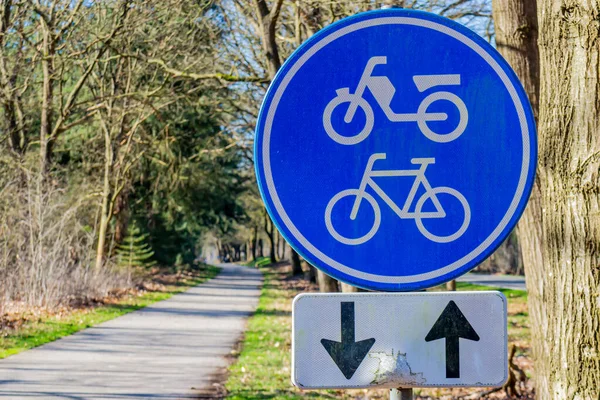 Trafik işareti: Her iki yönde bisiklet ve motosiklet şeridi, mavi ve beyaz yuvarlak plaka, Hollanda 'nın bulanık arka planında kaybolmuş ağaçlar arasındaki bisiklet yolu, güneşli bir gün