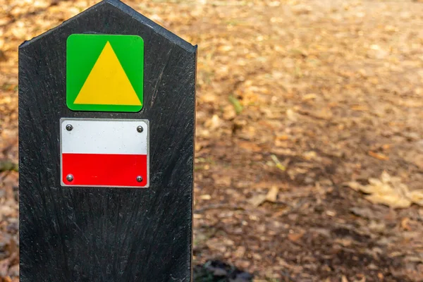Green with a yellow arrow, white and red lines, hiking trail signs, dry ground with some tree leaves in a blurred background, sunny day, Dutch nature reserve Natuurpoort Vennenhorst, Netherlands