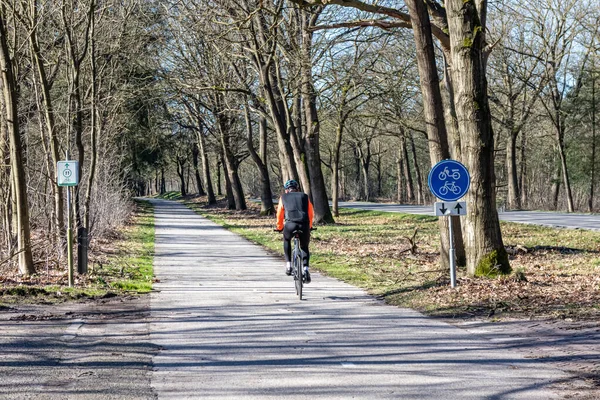Someren, Kuzey Brabant, Hollanda. 27 Şubat 2022. Bisikletçi bisiklet yolunda pedal çeviriyor, kırsal bir yolda, işaret veriyor: bisiklet ve moped yolu, rotalar 11, çıplak ağaçlar, kameraya dönüş, güneşli gün