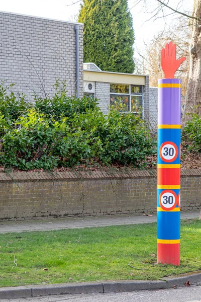 30 maximum speed sign in school zone, pole in red, blue and purple colors with a metal hand on top, school building in blurred background, Netherlands