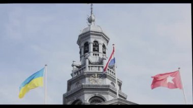 Ukrayna, Hollanda ve Maastricht bayrakları dalgalanıyor, belediye binasının kulesi, saat, çan ve rüzgar gülü, arkasında beyaz bulutlar olan mavi gökyüzü. Maastricht, Güney Limburg, Hollanda