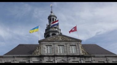 Belediye binası, Ukrayna bayrakları, Hollanda ve Maastricht dalgalanmaları, kulesi, saati, çanları ve rüzgar gülü, arkasında beyaz bulutlar olan mavi gökyüzü. Maastricht, Güney Limburg, Hollanda