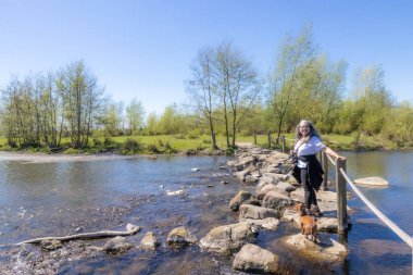 Brug Molenplas, basamak taşları, duruşu, gülümsemesi ve kamerasıyla olgun turist, sıradan kıyafetler, arka planda ağaçlar, Stevensweert, Güney Limburg, Hollanda 'da güneşli bir gün.
