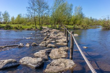 Oude Maas nehri, Brug Molenplas 'da köprü olarak kullanılan basamak taşları, ahşap direkler ve çitler, arka planda yeşil ağaçlar, Stevensweert, Güney Limburg, Hollanda' da güneşli bir gün.