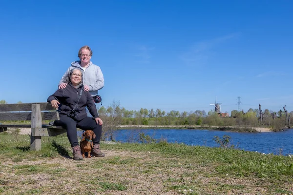 Olgun turist çifti, dachshund kameraları, Molenplas 'a bakış açısı arka planda doğa rezervi, nehir ve rüzgâr değirmeni, Stevensweert, Güney Limburg, Hollanda' da güneşli bir bahar günü