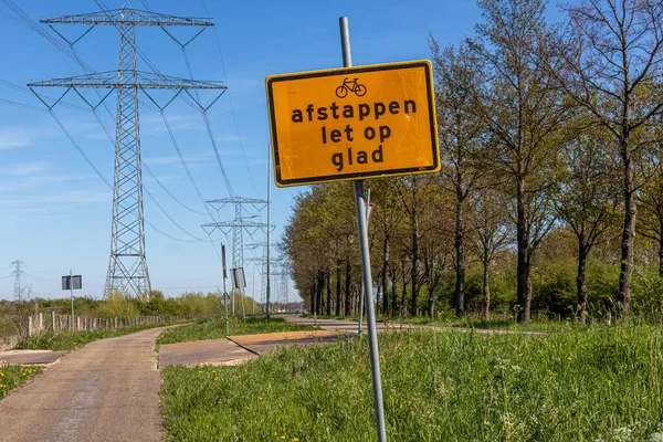 Metal sign with the Dutch inscription Afstappen, Let op, Glad, which means when getting off, watch out, slippery. Ciclovia, country road, trees and high voltage poles in the background, Netherlands