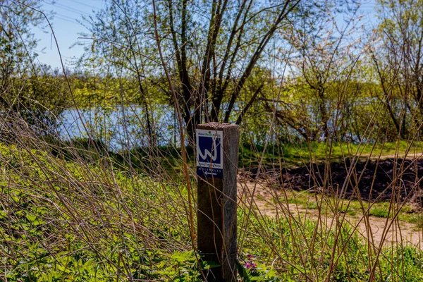 Wooden post with a hiking trail sign indicating the route to the Molenplas nature reserve, wild vegetation, Oude Maas river in the background, sunny day in Stevensweert, South Limburg, Netherlands