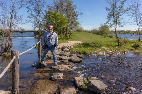 Olgun erkek yürüyüşçü Brug Molenplas 'ın basamaklarında duruyor, gülümsüyor ve kameraya bakıyor, günlük yürüyüş kıyafetleri, doğa koruma alanında güneşli bir gün, Stevensweert, Güney Limburg, Hollanda