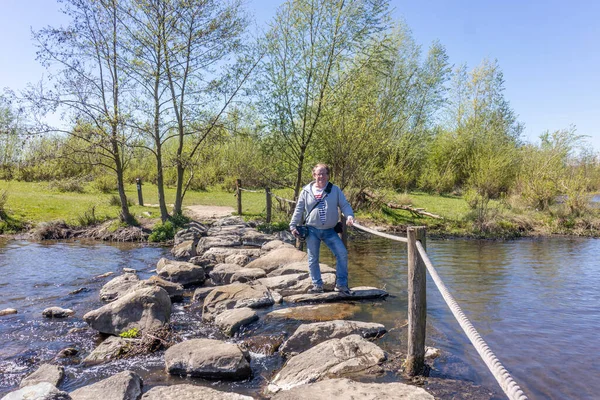 Brug Molenplas 'ın basamak taşları, kamerasıyla ayakta duran, gülümseyen ve kameraya bakan olgun erkek turist, günlük yürüyüş kıyafetleri, Stevensweert, Güney Limburg, Hollanda' da güneşli bir gün.